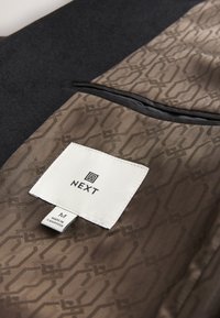 Gros plan de la doublure d'une veste marron à motifs avec une étiquette blanche de la marque NEXT et une étiquette taille M, montrant la poche intérieure et les détails du tissu.