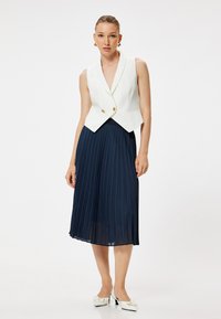 Witte mouwloze vest met gouden knopen, gecombineerd met een marineblauwe geplooide midi-jurk en witte hakken. Gladde stof en taillerende pasvorm.