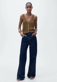 Gilet en tricot marron avec des boutons, associé à un jean en denim foncé à jambes larges. Texture lisse, design minimaliste et coupe décontractée. Des sandales complètent le look.