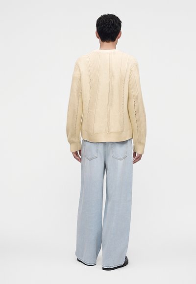 Homme portant un pull en maille crème et un jean large bleu clair, debout de dos sur un fond uni.