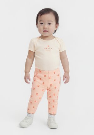 Toddler with short dark hair standing, wearing a cream "One of a Kind" shirt, peach floral pants, white socks, and white sneakers.