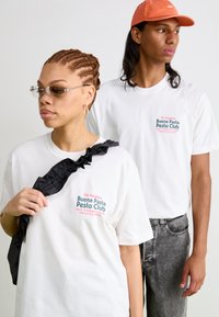 Two individuals wearing white t-shirts. One shirt features colorful text, with an accent black ruffled bag. The background is neutral.