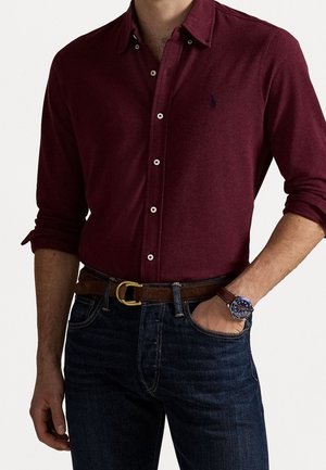 Hombre con camisa burdeos abotonada y mangas remangadas, jeans oscuros, cinturón marrón con hebilla dorada y reloj de pulsera con esfera azul.