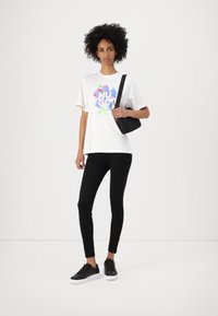 Tricou grafic alb cu text colorat "HUGO", purtat cu colanți negri și adidași negri. Modelul ține o mică geantă de mână neagră.