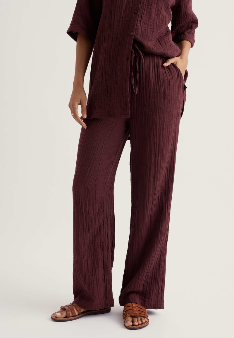 Personne portant un pantalon ample et texturé de couleur bordeaux foncé avec un haut boutonné assorti et des sandales plates marron à bout ouvert, une main dans la poche.