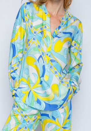 Kvinde iført livlig blå, gul og grøn abstrakt mønstret skjorte med knapper og matchende bukser med hænderne i lommerne.