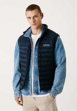 Marinblå puffer-väst över en ljusblå denimjacka, med dragkedja fram, huva och Red Bull Racing-logotyp på bröstet. Beiga byxor.