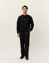 Svart sweatshirt med rund halsringning og subtil logo, kombinert med løstsittende svarte bukser. Modellen har på seg brune lær-sko.