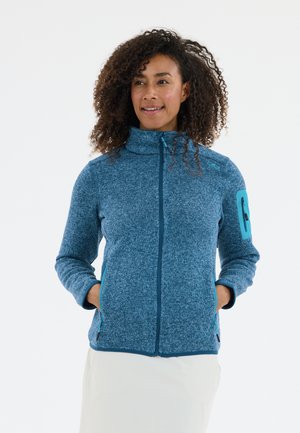 Kvinde med krøllet hår iført en blå fleecejakke med lynlås og lynlåslommer samt en lynlåslomme på ærmet, hænderne i lommerne, let smilende.