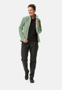 Giacca impermeabile verde chiaro con chiusura a zip, abbinata a pantaloni impermeabili neri e robuste scarpe nere. Texture liscia e design aderente.