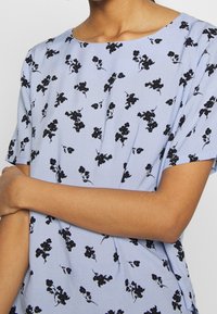 Světle modrý krátký rukáv s kulatým výstřihem, zdobený černým květinovým vzorem po celém povrchu a volným střihem. Měkká, texturovaná látka.