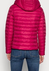 Tikkitakki fuksianpunaisena, jossa on vaakasuora tikkaus, huppu ja sileä, kiiltävä pinta. Yhdistetty istuviin sinisiin farkkuihin.