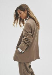 Blazer marrón a medida y pantalones a juego, con solapas anchas, bolsillos laterales y una textura suave, llevados sobre una camisa blanca.