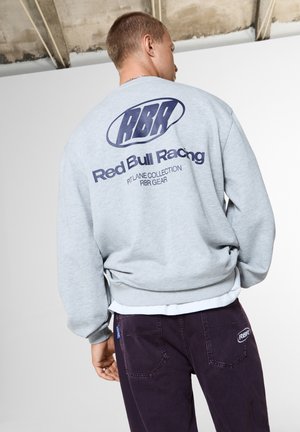 Мъж, носещ светлосива суитчър Red Bull Racing и тъмни панталони с лого на RBR на задните джобове, стои на закрито срещу бяла стена.