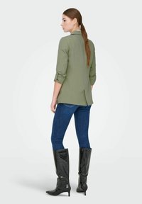 Blazer sur mesure vert olive, manches retroussées et une fente arrière, associé à un jean bleu foncé ajusté et des bottes noires brillantes montant jusqu'aux genoux.