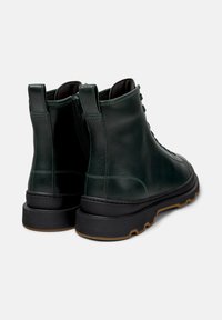 Botas de tobillo de cuero verde oscuro con una textura suave, suelas negras y cordones decorativos. Cuentan con una cremallera lateral para facilitar su uso.