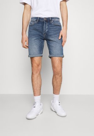 Hombre vistiendo pantalones cortos de mezclilla azul arremangados, calcetas blancas, zapatillas blancas y una camiseta blanca, de pie contra un fondo claro y liso.