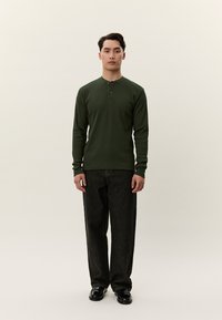 Fiatal férfi áll, sötétzöld hosszú ujjú henley pólót, laza fekete farmert és fekete cipőt visel, egyszínű háttér előtt.