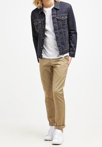 Giacca di jeans blu scuro con dettagli di cucitura visibili, t-shirt bianca sotto, chinos beige e sneakers bianche.