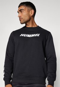 Sort sweatshirt med rund halsudskæring, ribbede manchetter, og et stort hvidt "HUMMEL" logo på brystet. Fremstillet af blødt materiale.
