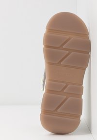 Semelle d'une basket en caoutchouc beige avec un motif géométrique de la bande de roulement et un logo en relief discret sur une surface blanche.