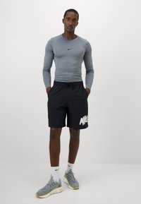 Vyras vilkintis pilką ilgomis rankovėmis sportinę maikę, juodas Nike šortus su balta logo, baltas Nike kojines ir pilkas bėgimo batus su geltonais padais stovi.