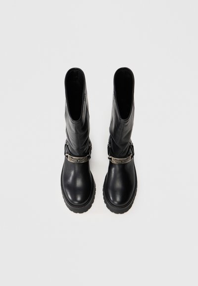 Botas de cuero negro hasta la rodilla con puntera redonda, cuerpo texturizado y un acento decorativo de hebilla plateada alrededor del tobillo. Suela de goma.