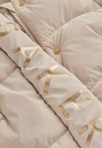 Doudoune beige avec une inscription et des boutons dorés. Présente une texture matelassée avec une finition lisse et une fermeture frontale zippée.