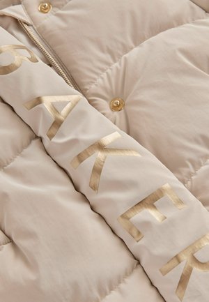 Doudoune beige avec une inscription et des boutons dorés. Présente une texture matelassée avec une finition lisse et une fermeture frontale zippée.