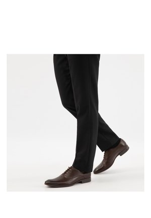 Beine in schwarzen Anzughosen, die polierte dunkelbraune, aus Leder gefertigte Schnürschuhe tragen und vor einem schlichten weißen Hintergrund gehen.