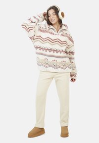 Przytulny, oversize polarowy pulower w odcieniu kremowym z dzianinowymi geometrycznymi wzorami w kolorach czerwonym, niebieskim i złotym; zestawiony z kremowymi spodniami i beżowymi kapciami.
