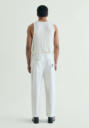 Hombre de pie de espaldas vistiendo una camiseta blanca sin mangas, pantalones blancos con bordado en los bolsillos y zapatos negros sobre un fondo liso.