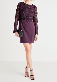 Lila klänning med långa spetsärmar, texturerad bodice och samlad midja; tillsammans med en svart clutchtaske och svarta klackskor med remmar.