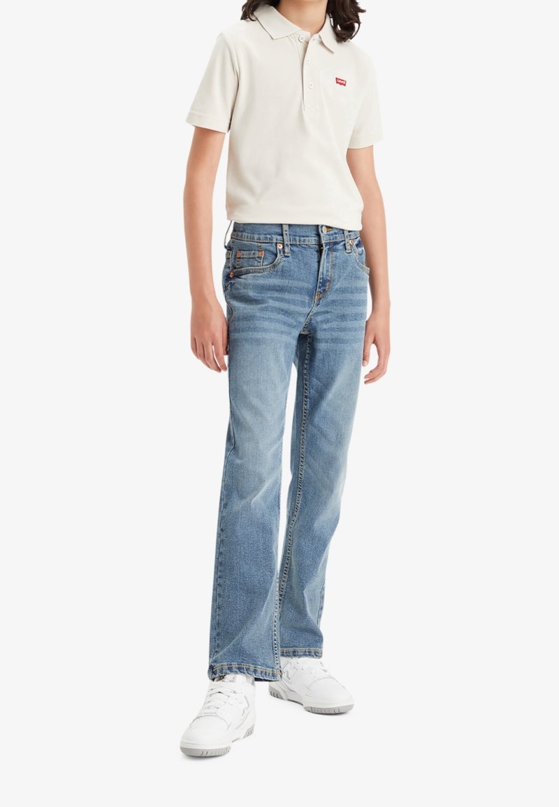 Camisa polo beige con cuello, pantalones vaqueros de mezclilla azul con un lavado claro y zapatillas blancas, de pie con las manos relajadas a los lados.