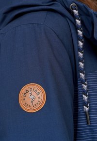 Marinblå jacka med en rund beige läderpatch med en flamingotavla och flätad dragsko med svarta, vita och blå accenter.