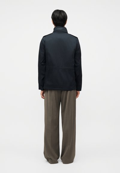 Personne debout, vue de dos, portant une veste noire et un pantalon marron ample, sur un fond clair uni.