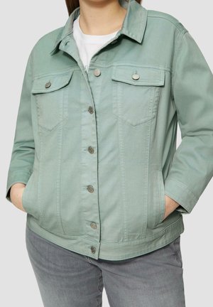 Personne portant une veste vert clair boutonnée avec des poches à rabat et les mains dans les poches latérales, associée à un pantalon gris et une chemise blanche.