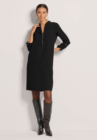 Schwarzes Kleid mit einem Reißverschlusskragen, langen Ärmeln, die an den Bündchen hochgerollt sind, und einer Brusttasche; kombiniert mit knielangen braunen Lederstiefeln.