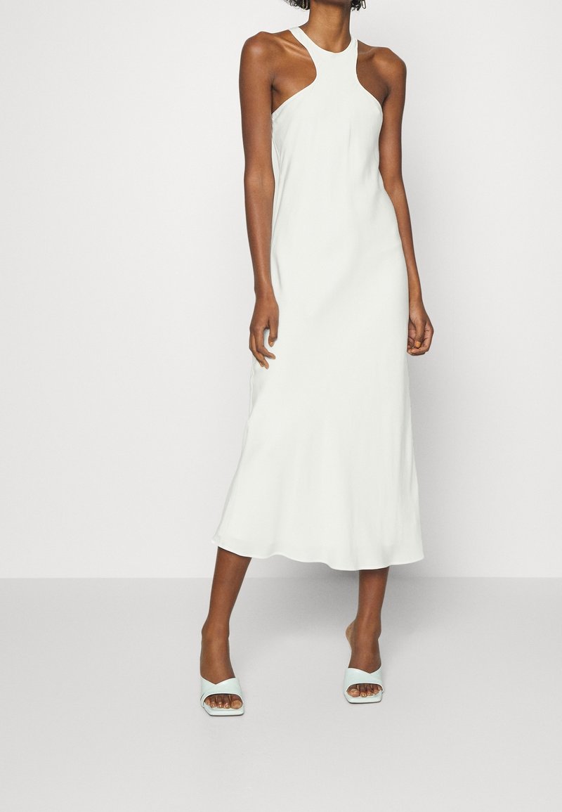 Femme portant une robe midi blanche sans manches avec un décolleté asymétrique et des talons blancs ouverts, debout sur un fond uni.