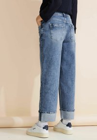 Vaqueros de mezclilla celeste con un corte relajado, puños doblados y bolsillos traseros. Combinados con zapatillas blancas con detalles en azul marino.