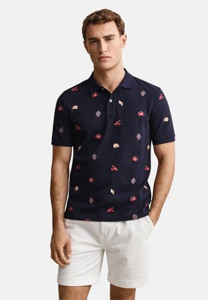 Homme portant un polo bleu marine avec des motifs rouges de crabes et de coquillages, porté avec un short blanc, debout avec une main dans la poche.