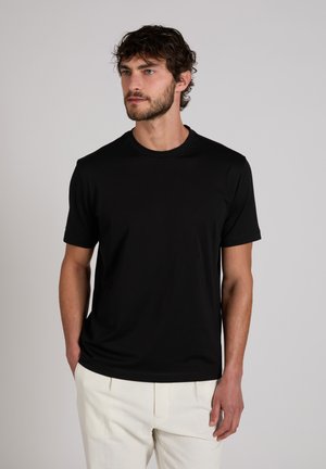 Hombre con cabello corto y rizado y barba, vestido con una camiseta negra de manga corta y pantalones blancos, de pie con una mano en el bolsillo.
