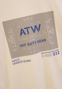 Creme bomuld t-shirt med et sølvfarvet grafisk print og blå tekst. Designet inkluderer tekniske elementer og forskellige tal.