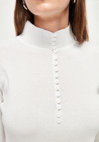 Col roulé blanc à côtes avec une rangée de 11 petits boutons blancs sur le devant. Texture lisse et coupe ajustée pour une apparence élégante.