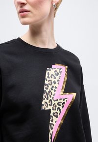 Κοντινό πλάνο ατόμου που φοράει μαύρο φούτερ με σχέδιο λεοπάρ, ροζ και χρυσή αστραπή, και ασημένιο κρίκο σκουλαρίκι στο αριστερό αυτί.