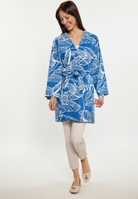 Abito avvolgente blu con motivo floreale, maniche lunghe, vita con cintura e vestibilità ampia, abbinato a pantaloni beige chiari e scarpe.
