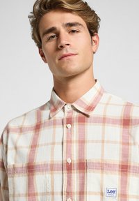 Camisa de cuadros de manga corta con una tela suave y ligera en colores crema, melocotón y marrón claro; presenta un cuello abotonado y una etiqueta con el logo.
