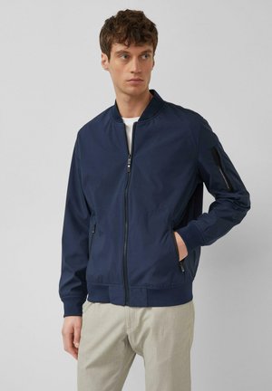 Jeune homme portant une veste bomber bleu marine avec fermeture éclair, poche sur la manche gauche, et un pantalon beige clair, debout devant un fond uni.