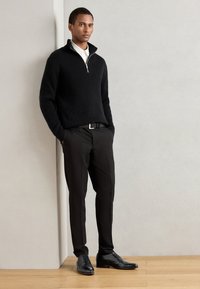 Pullover nero a coste con colletto a mezze zip, pantaloni neri e scarpe nere con texture. Sfondo neutro e pavimento in legno liscio.