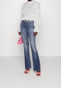 Blouse blanche à manches longues avec un col à volants, associée à un jean bleu évasé. Accessoires comprenant un petit sac à main rose et des sandales à talons roses.